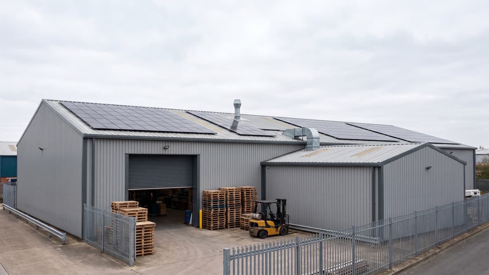 An industrial unit with solar panels across its shallow-pitch steel roof above a concrete yard.
