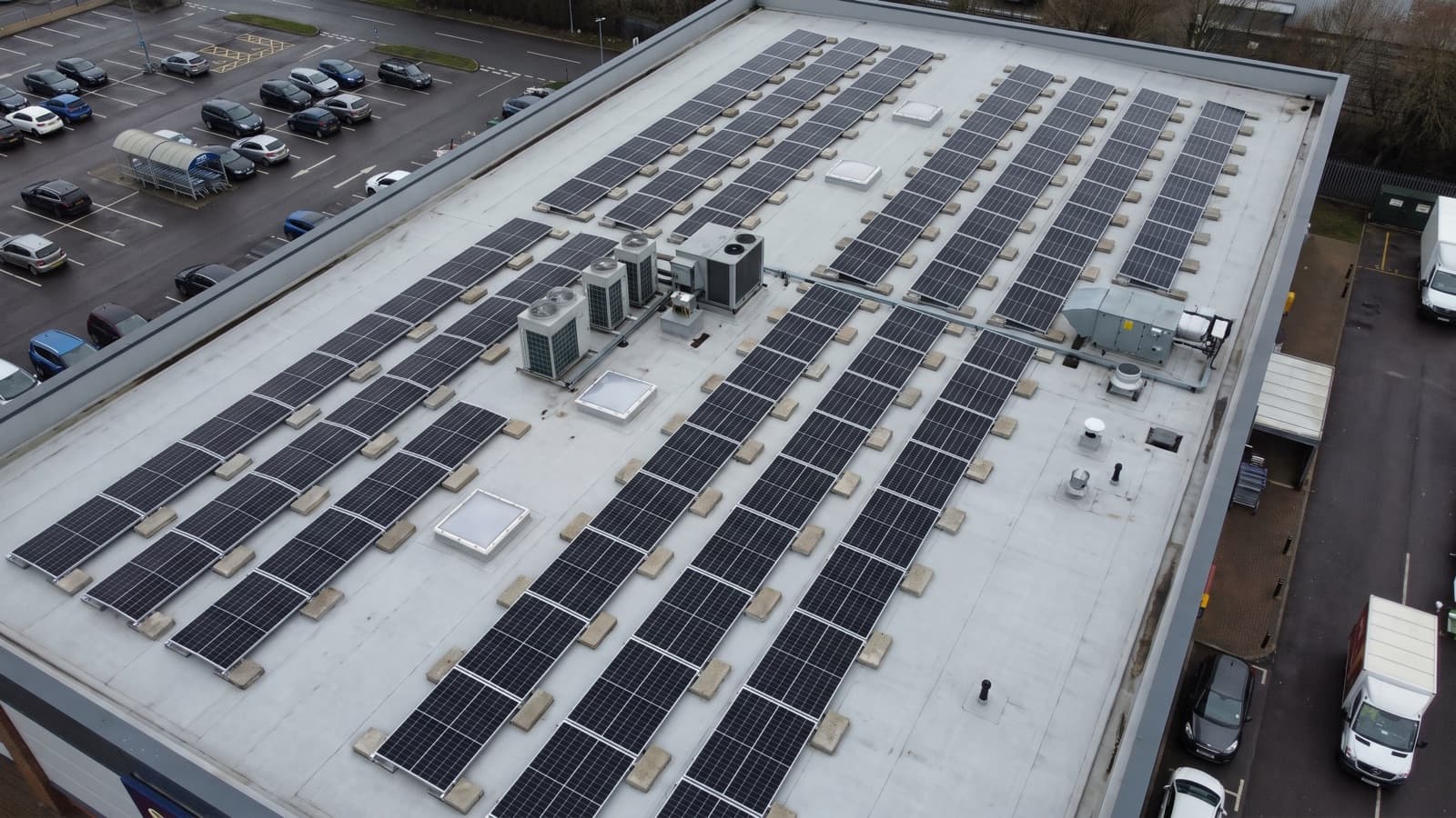 Aerial view of a retail park unit's flat roof carrying a ballasted solar array in rows between rooftop plant.