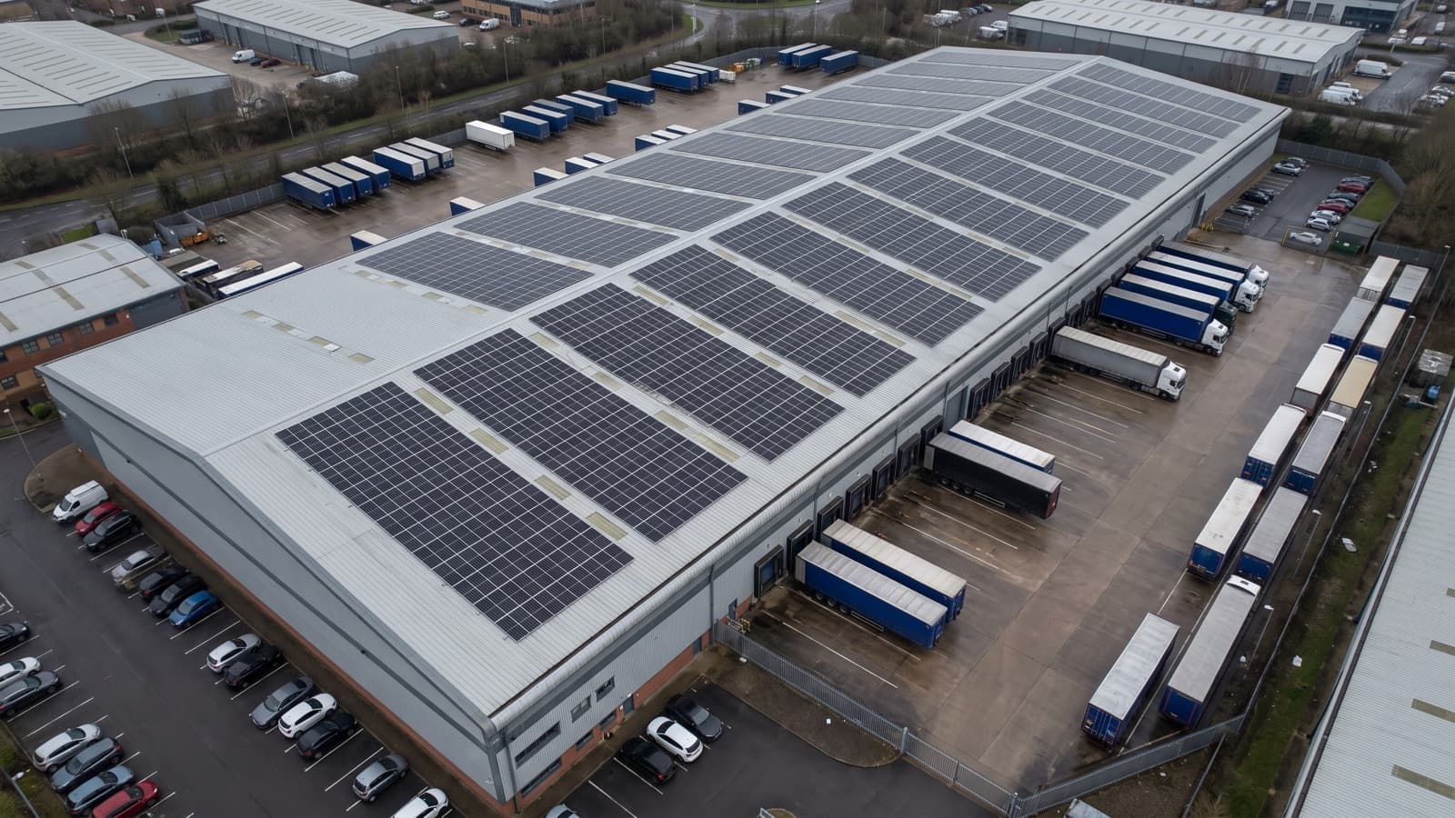 Aerial view of a distribution warehouse with a large solar array across its steel roof and trailers at the loading docks.